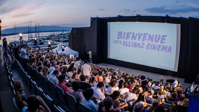 Un grand écran de cinéma en plein air est installé au bord du lac Léman, avec une foule de spectateurs assis en gradins. La projection a lieu à la tombée de la nuit, sous un ciel violet orangé avec la lune brillante. En arrière-plan, on aperçoit le lac, des arbres et un bâtiment éclairé.