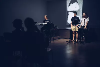 Spoken Worlds: L’Enfant hors champ Photo d'une lecture sonore avec un musicien assis avec appareils électroniques et deux femmes debout lisant à un micro.
