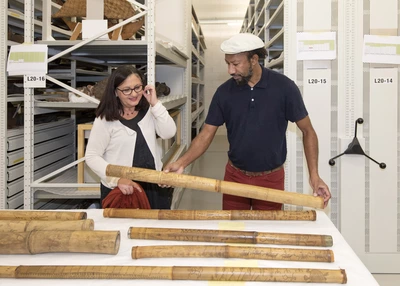 Visite "Encounters" Sur cette image, Denis Pourawa, un poète et écrivain kanak (Nouvelle-Calédonie), est dans la réserve des collections du MEG avec la conservatrice des collections Océanie. Denis Pourawa a devant lui une série de bambou gravé kanak. Il en prend un dans les mains et pose son regard dessus. Les bambous kanak mesure environ 1m20 de long.