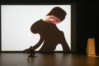 Une personne sur scène dont l'image est projetée sur le fond de scène, l'image est fortement déformée.