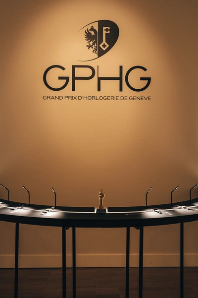 L’image montre un mur beige avec le logo du GPHG – Grand Prix d’Horlogerie de Genève inscrit en grand noir. Au centre, sous le logo, se trouve une vitrine semi-circulaire noire avec un éclairage discret, présentant des montres de prestige. Au milieu de cette vitrine trône un petit trophée doré en forme de main levée. L’ambiance est élégante, sobre et muséale, mettant en valeur l’excellence de l’horlogerie.
