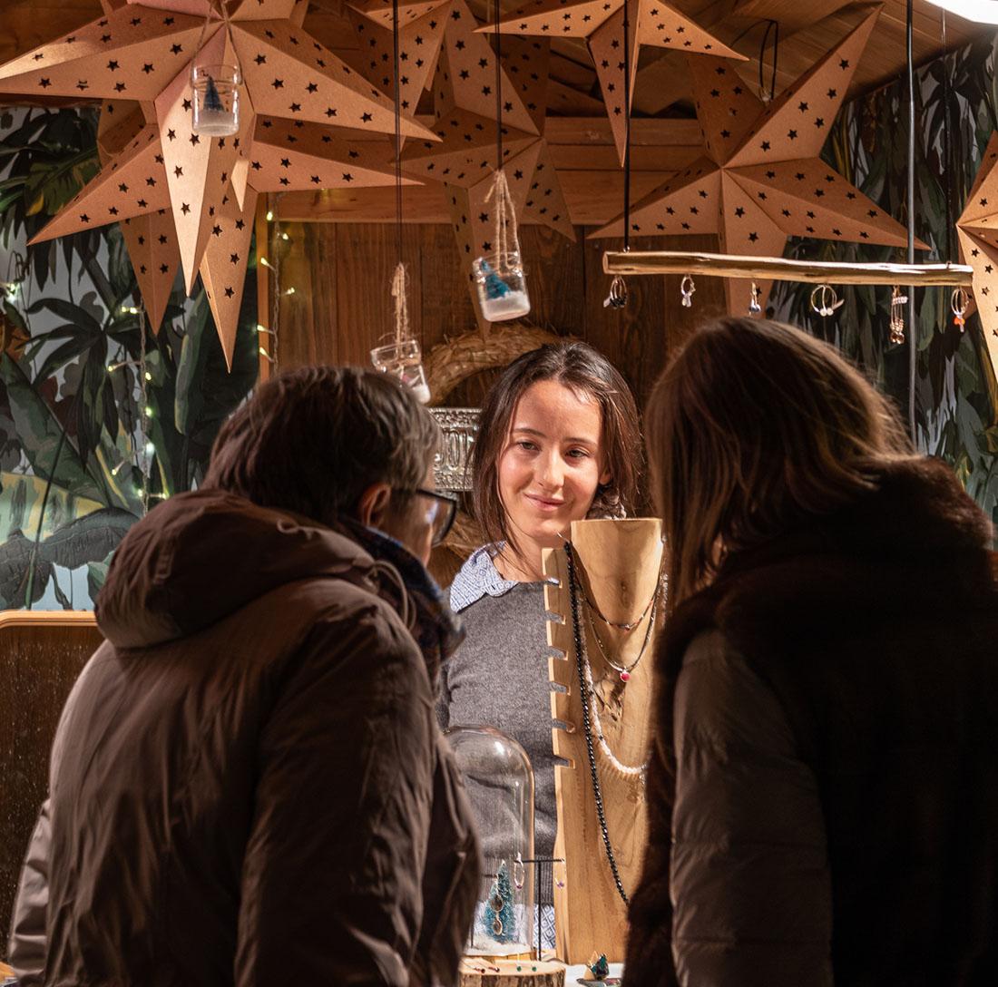 Besucher wählen handgemachten Schmuck bei einer Kunsthandwerkerin in einem mit Papiersternen geschmückten Weihnachtsmarkt-Chalet aus Holz