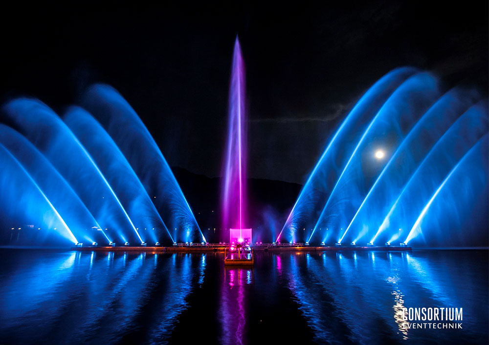 Illuminated fountain jets arcing over Lake Geneva at night in blue and violet light, with the moon in the sky