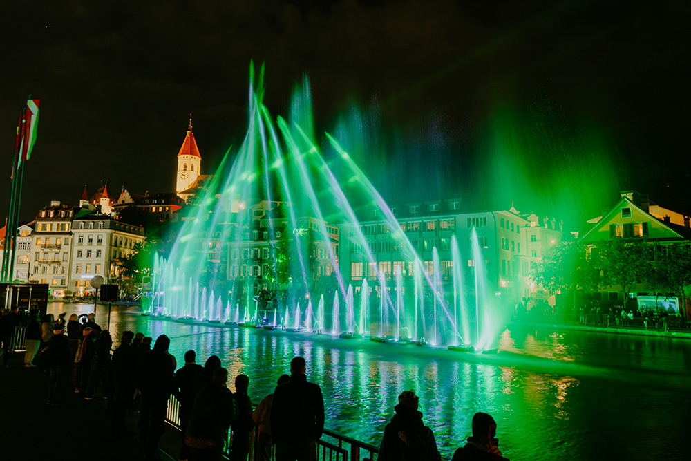 Illuminated fountains and lasers over a lake at night during a water and light show, viewed by a crowd on the shore